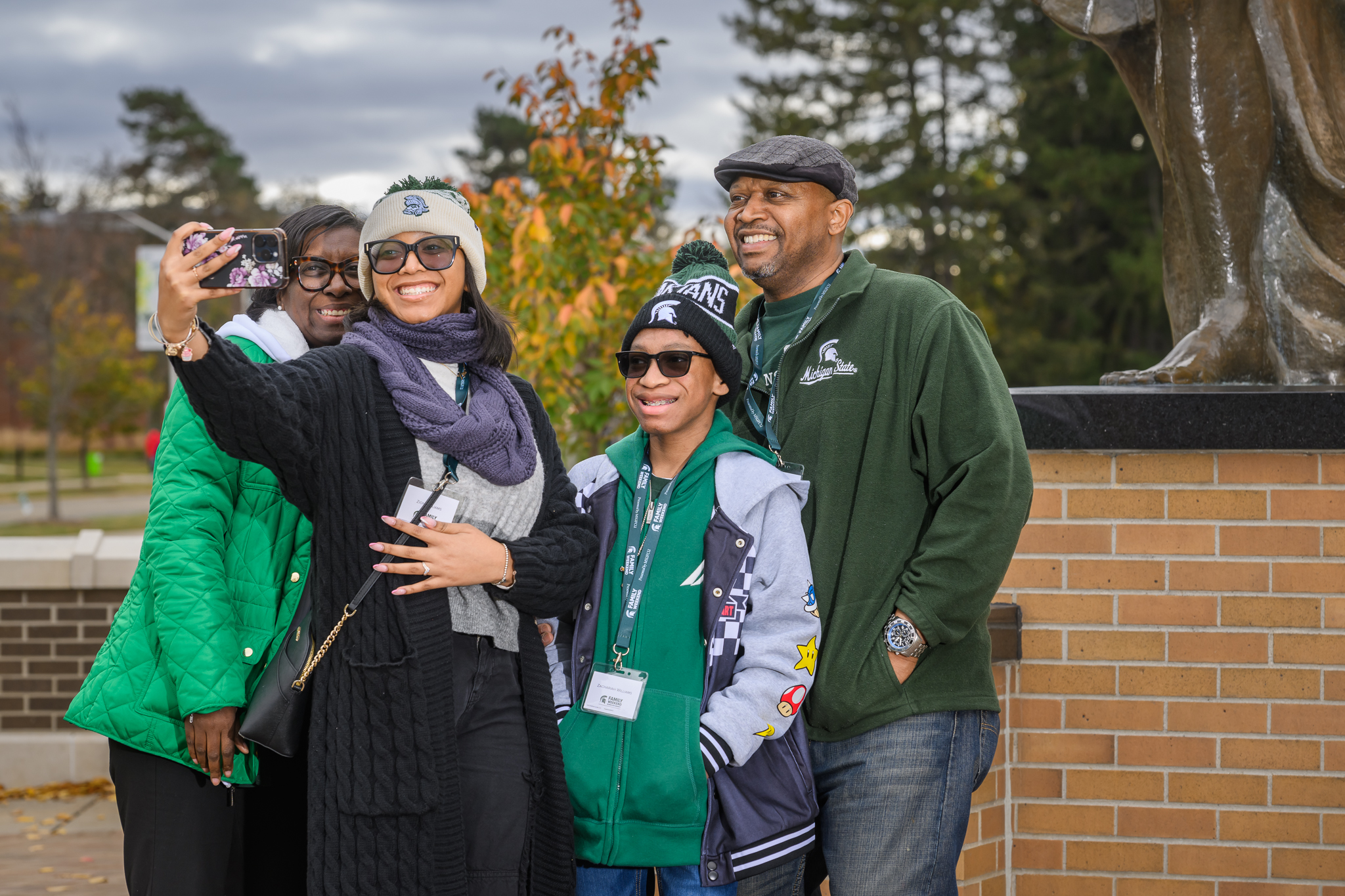 A smiling group takes a selfie in front of The Spartan statue at Michigan State University during a campus event, wearing green and white apparel to show Spartan pride.