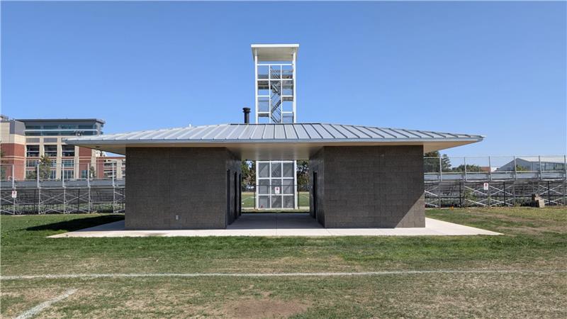 Small stadium support building with a metal roof and open center walkway, set beside athletic fields and bleachers under a clear blue sky.