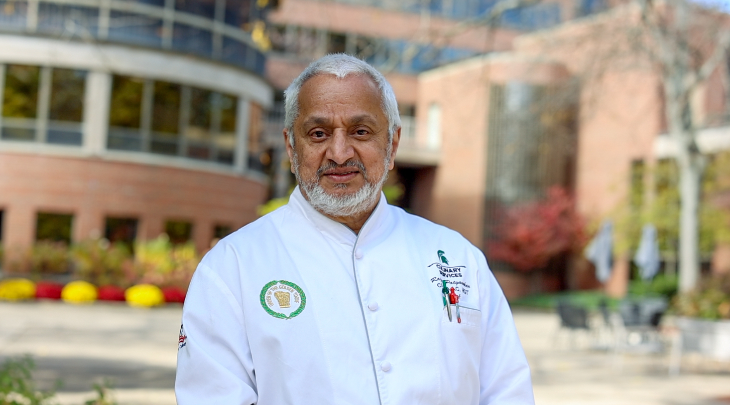 Chef Rajeev wearing a white Culinary Services jacket stands outdoors on Michigan State University’s campus, with fall foliage and brick buildings in the background.