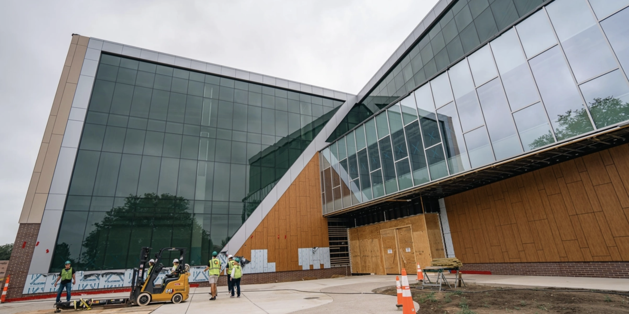 Modern recreation center under construction with large glass walls, wood panels, and construction workers using a forklift outside the entrance.