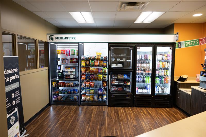 Convenience food items including prepackaged meals, soft drinks and bottled water on display in coolers under a Michigan State logo.