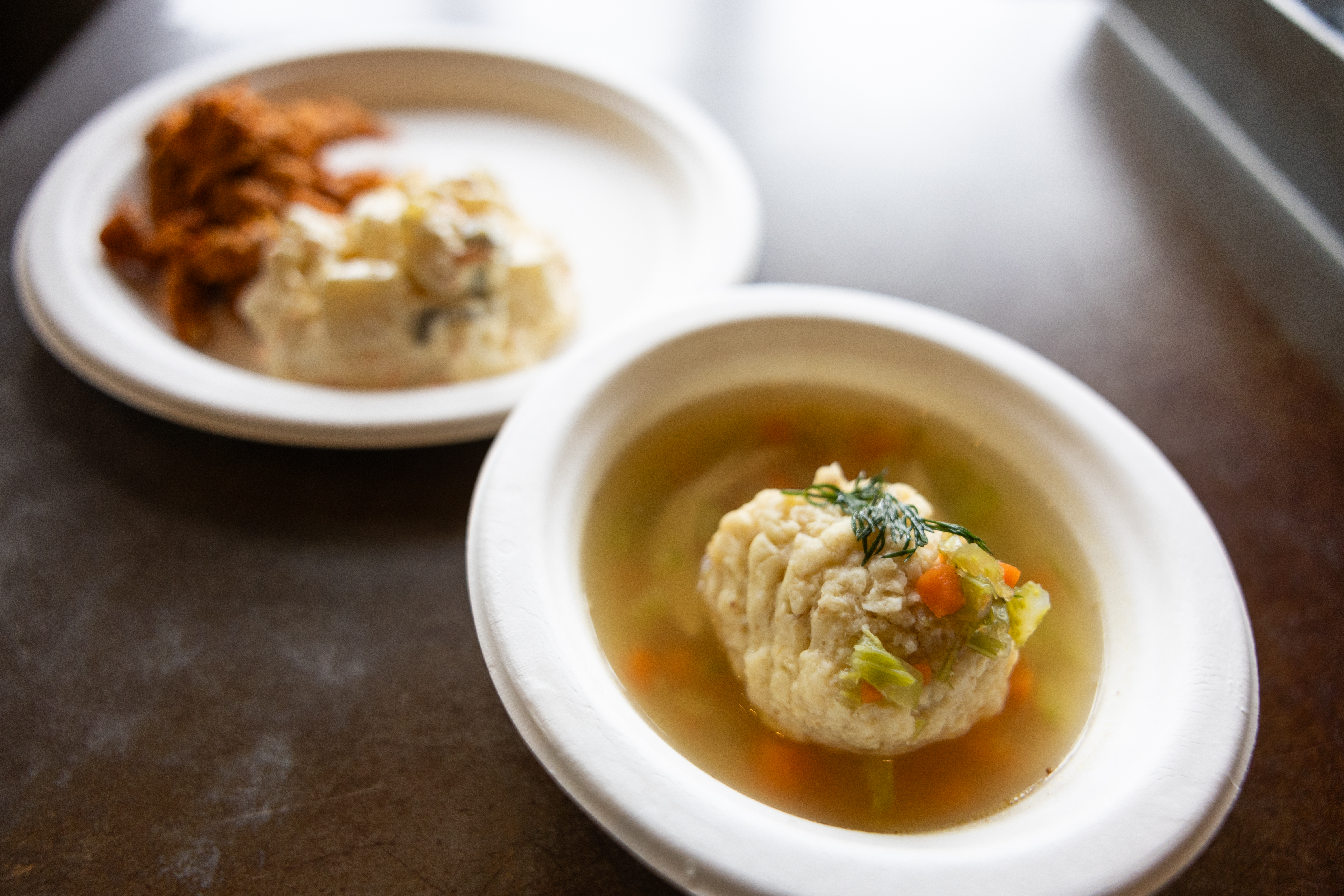 Bowl of clear soup with a large matzo ball and vegetables, with a plate of side dishes blurred in the background.