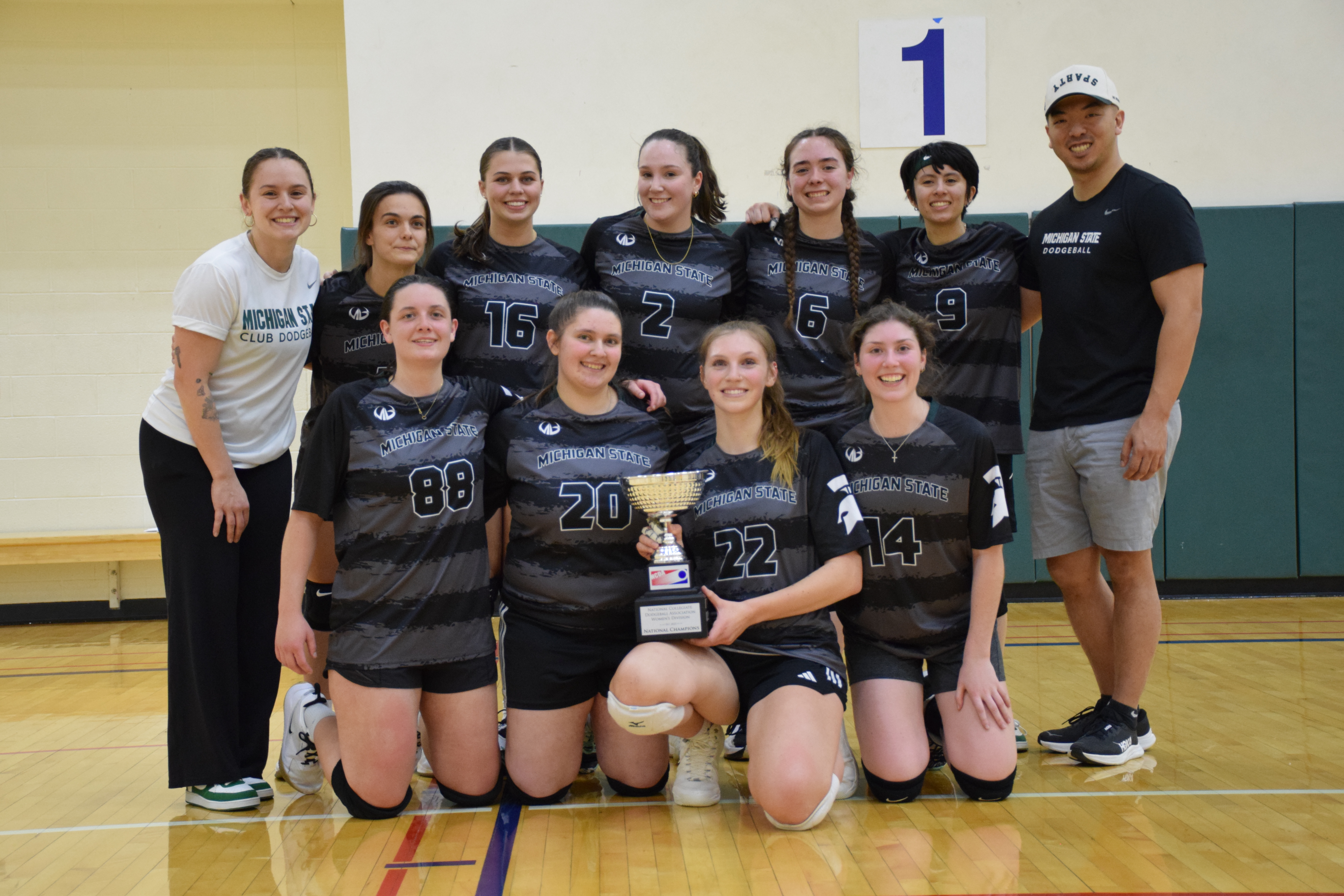Members of the MSU women's club dodgeball team pose on a court with their two coaches and holding a trophy after winning the national title. 