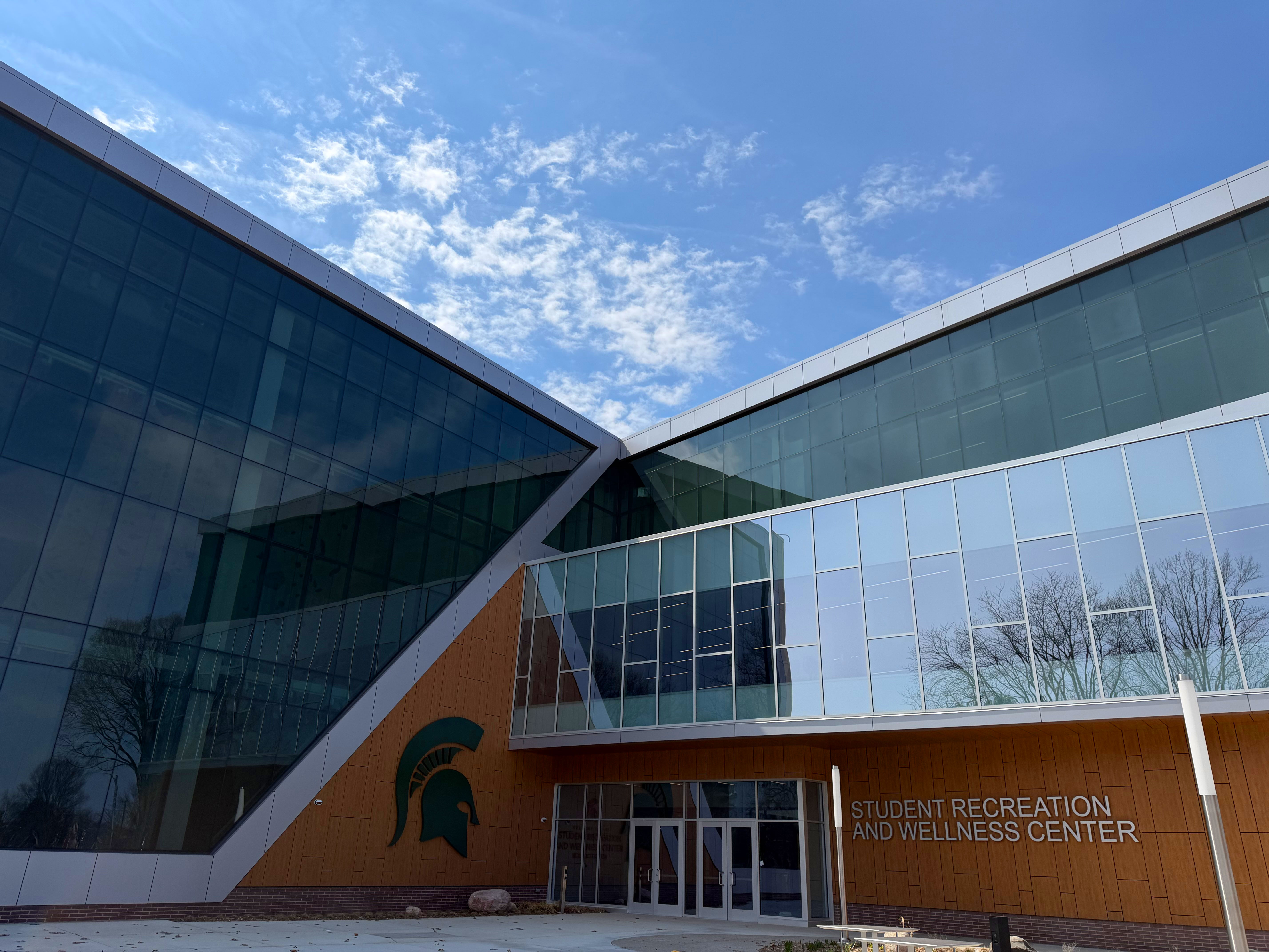 The front entrance of the new Student Recreation and Wellness Center on MSU’s campus, large glass window panes surround a Spartan helmet at the front doors.