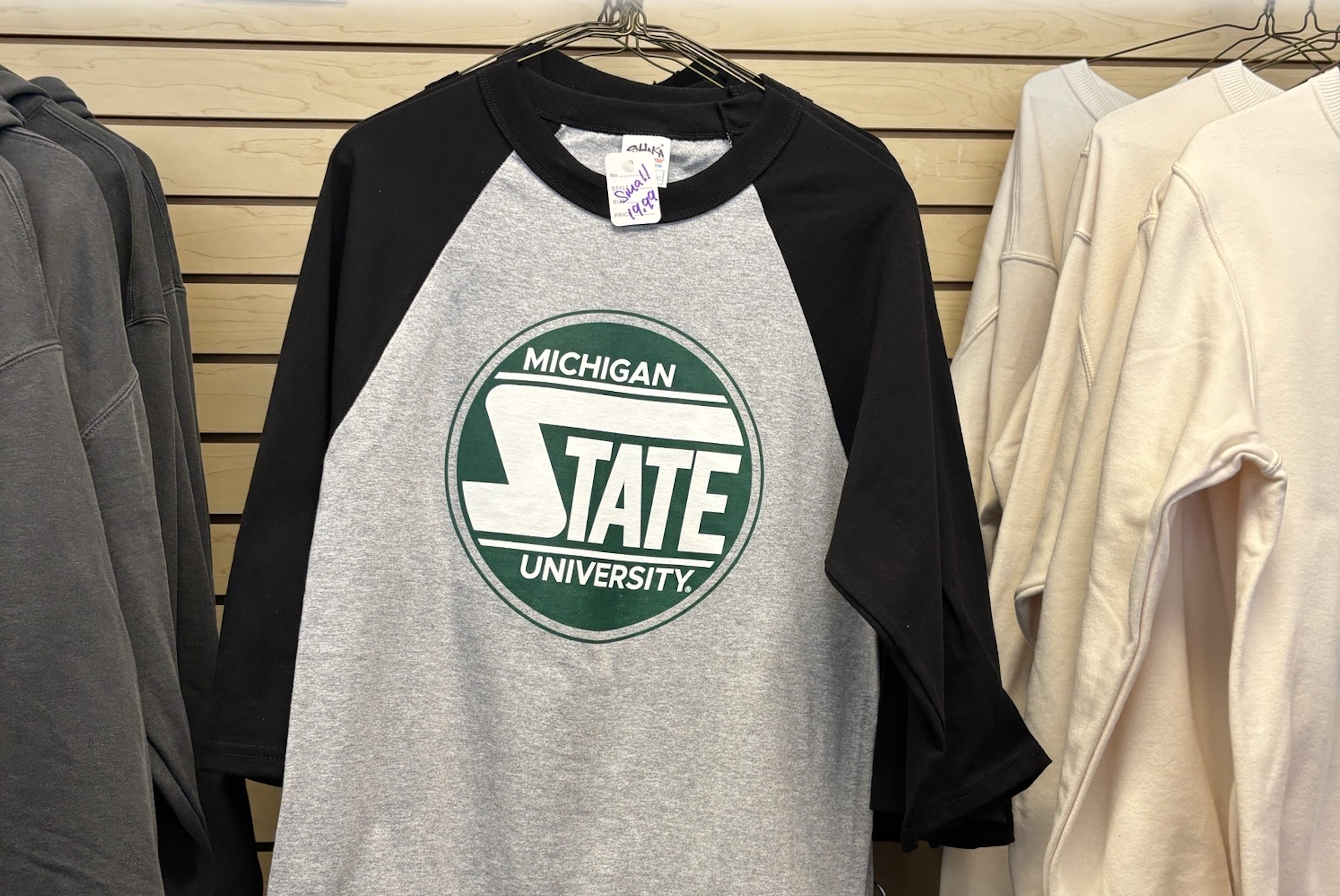 A black and gray baseball-style shirt with unique circular MSU graphic on front hangs for sale at Spartan Linen Services.