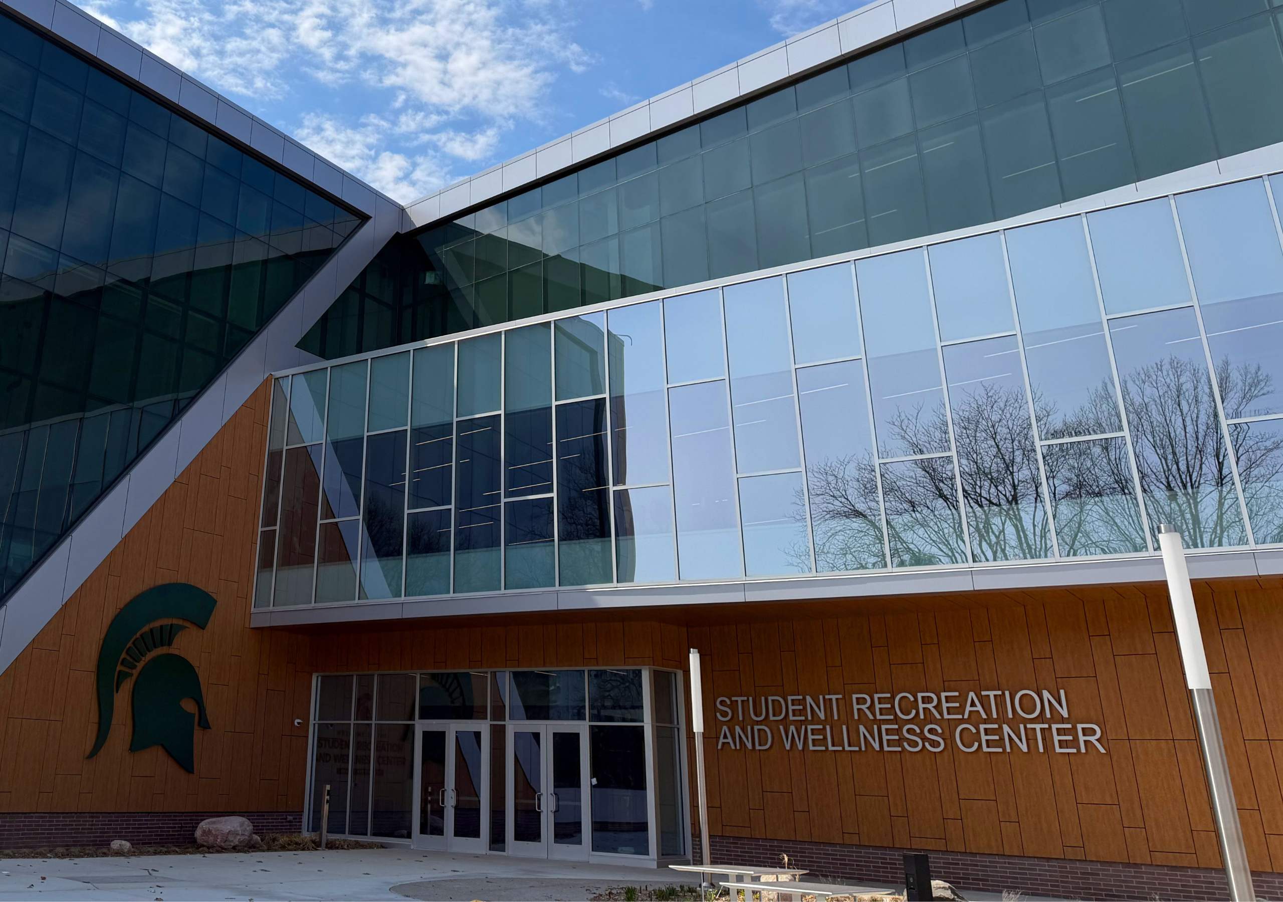 Modern glass and wood building exterior with large windows and angular architecture, featuring a Michigan State Spartan logo and the sign “Student Recreation and Wellness Center” near the entrance under a bright blue sky.