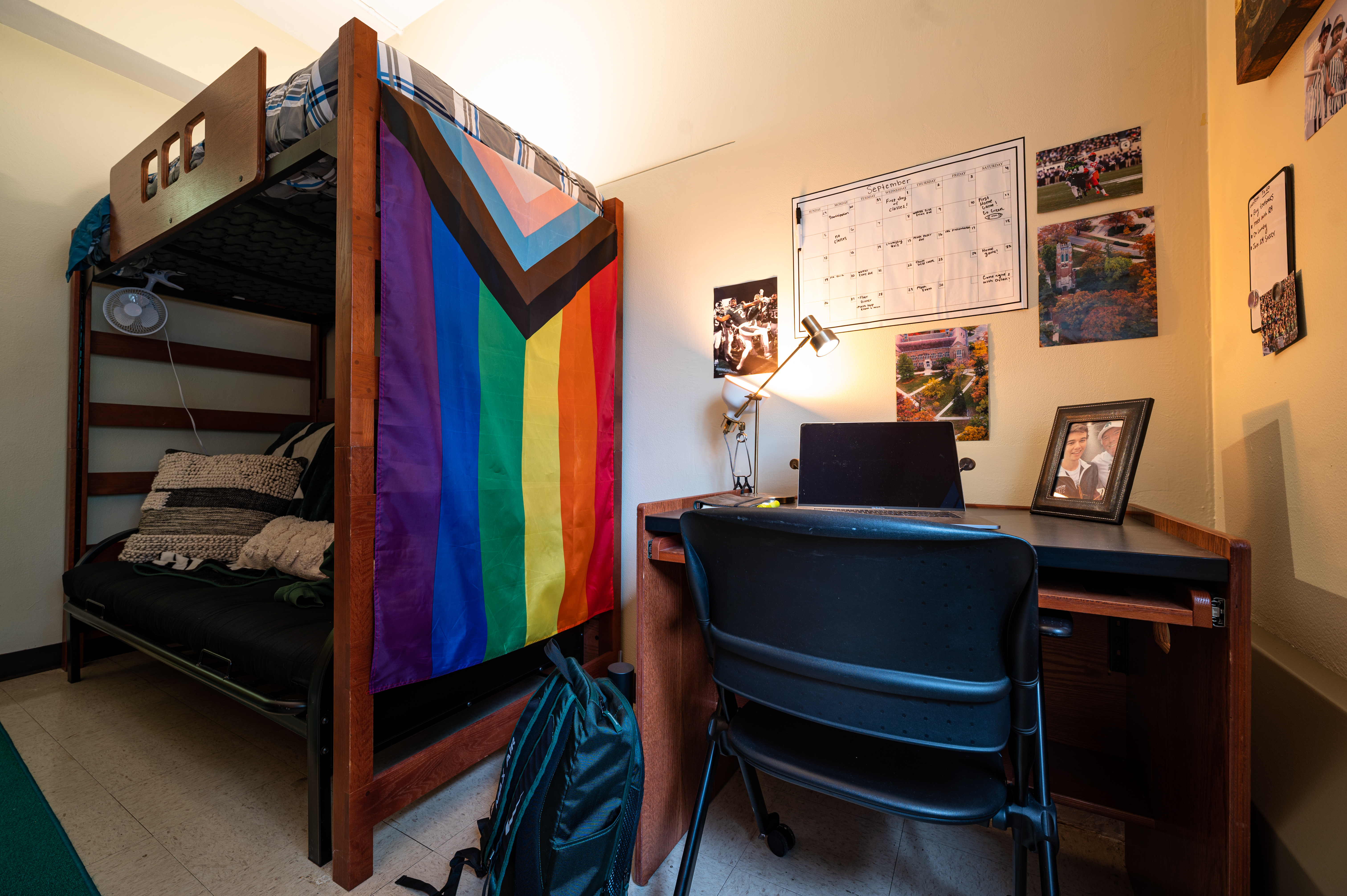 A warmly lit college dorm room with a wooden lofted bunk bed on the left and a desk on the right. A large Progress Pride flag hangs from the side of the bed above a cushioned futon. The desk holds an open laptop, adjustable lamp, and framed photo, with a black chair in front and a backpack on the floor. A monthly calendar, campus photographs, and other personal pictures decorate the walls.
