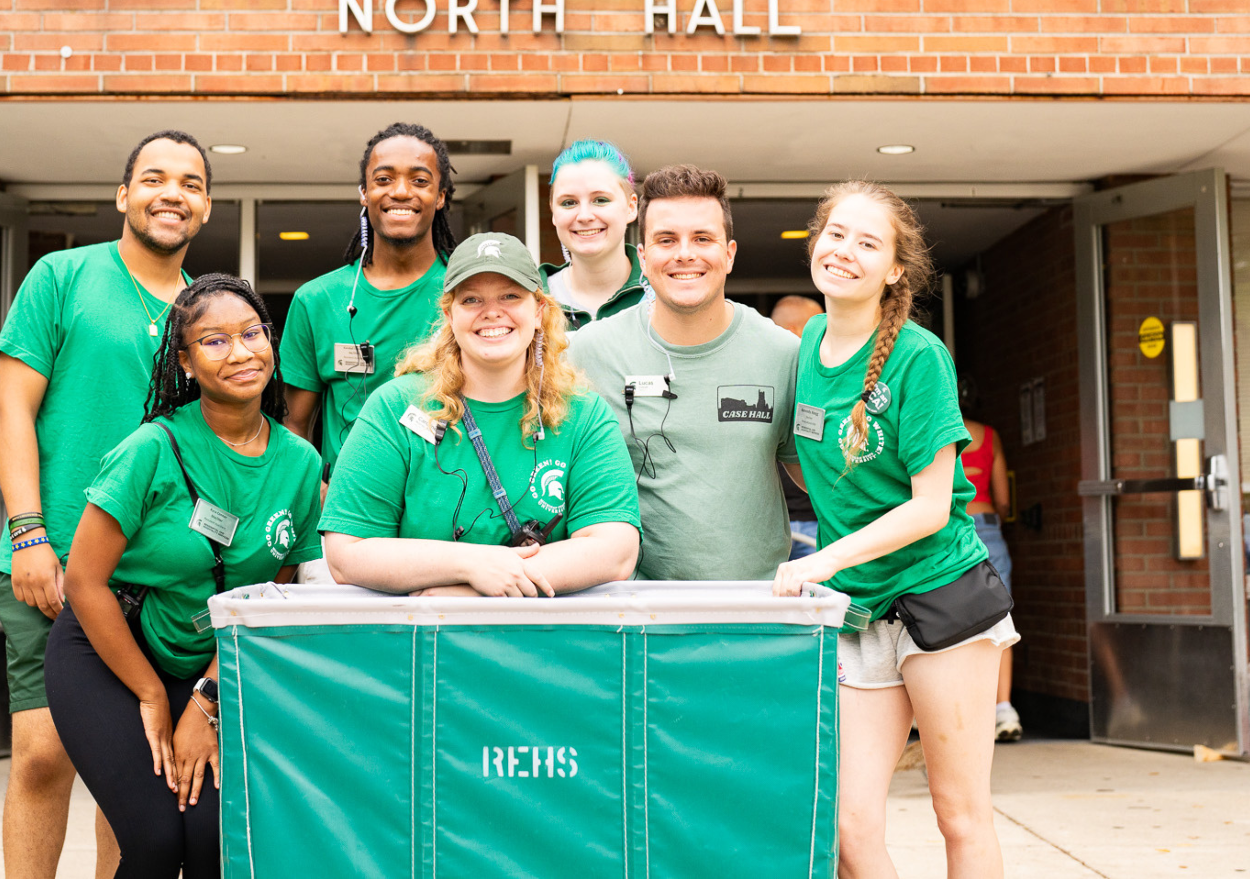 Seven people wearing green shirts pose together behind a green move-in cart outside Albert H. & Sarah A. Case North Hall. Additional carts and move-in supplies are visible near the residence hall entrance.