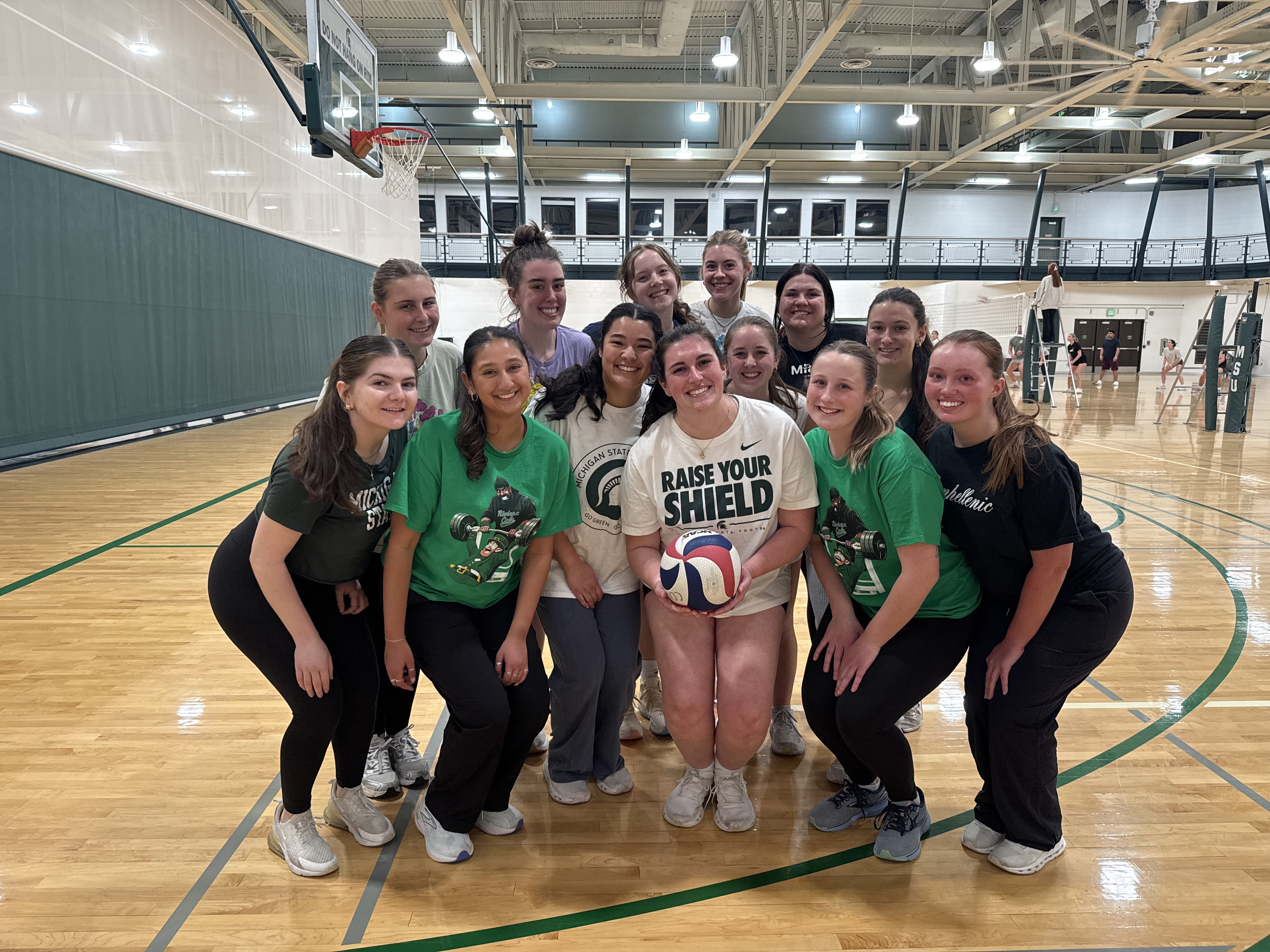 A group of 13 people pose together on a polished indoor volleyball court, smiling toward the camera. The person in the center holds a red, white, and blue volleyball and wears a “Raise Your Shield” T-shirt. Several others wear green Michigan State–themed shirts. Volleyball nets, basketball hoops, and additional players are visible throughout the brightly lit gym.