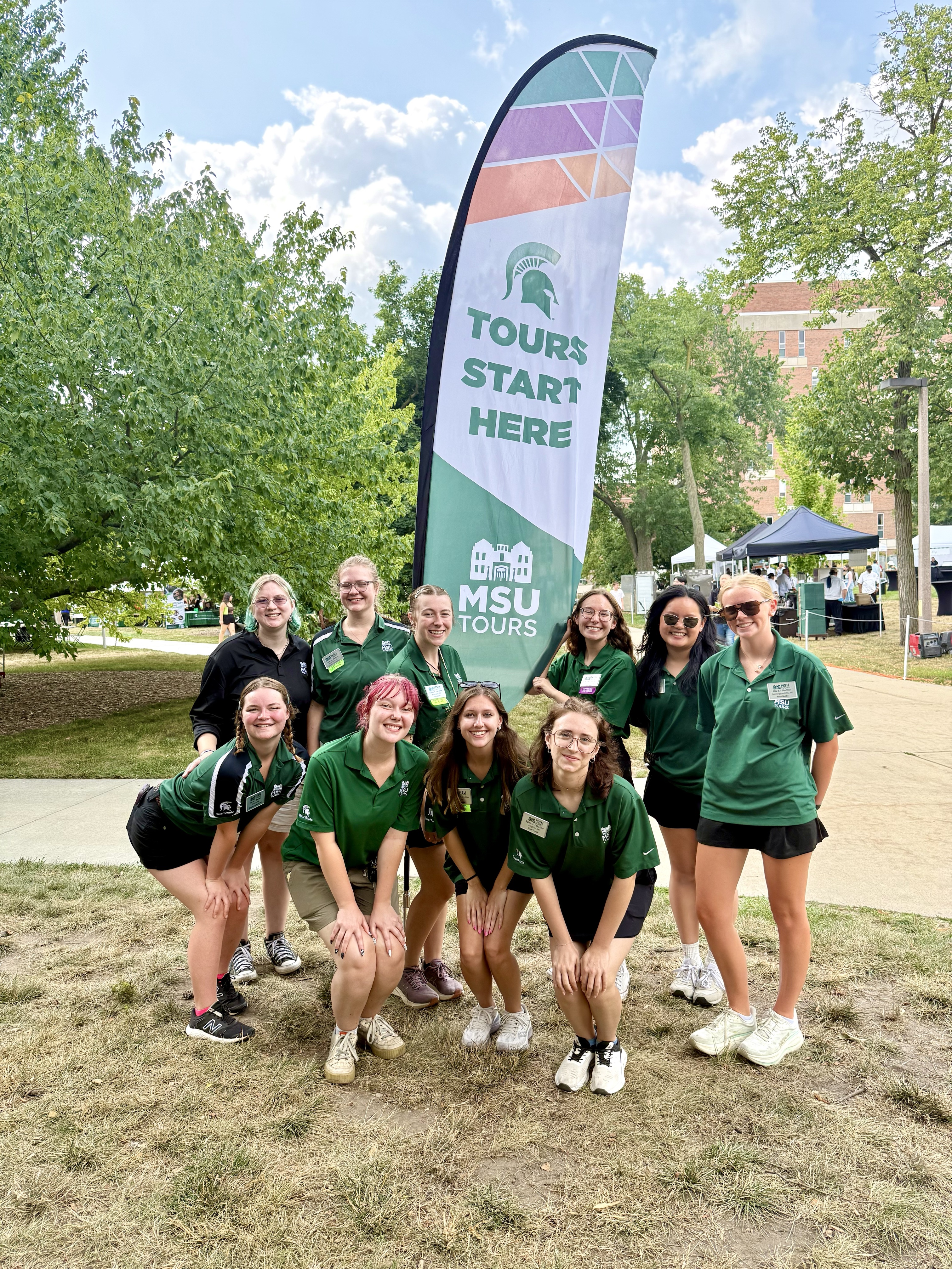 An MSU Tours group poses under a Green and White Days Tours Start Here banner on campus.