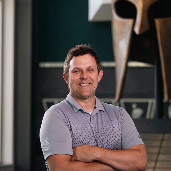 Professional headshot of a Michigan State University staff member in a gray collared shirt, smiling with arms crossed in front of a Spartan helmet sculpture.