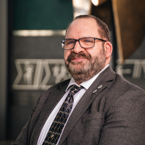 Bearded man wearing glasses, a suit jacket and tie, seated indoors and smiling slightly in a professional portrait setting.