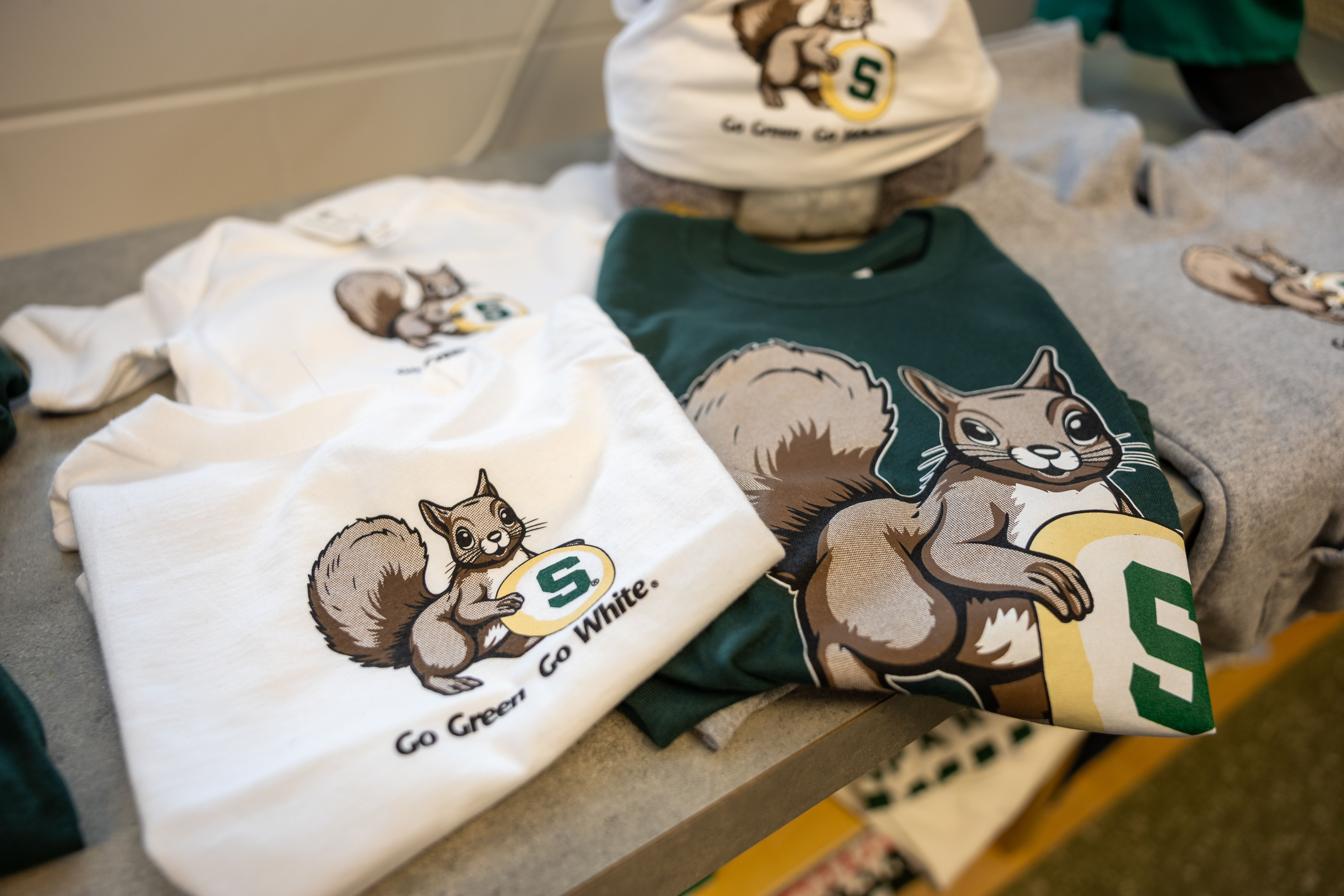 Two folded T-shirts at Spartan Linen Shop feature cartoon squirrels holding an MSU Bakers sugar cookie above the copy: Go Green Go White.