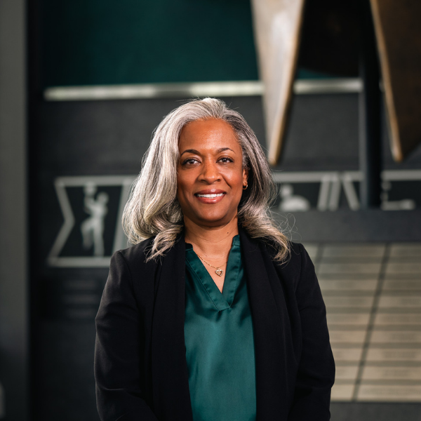Professional headshot of a Michigan State University staff member with long gray hair, wearing a green blouse and black blazer, standing in front of a Spartan helmet sculpture