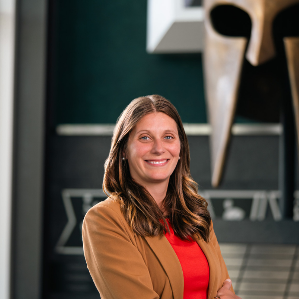 Professional headshot of a Michigan State University staff member in a red blouse and tan blazer, smiling in front of a Spartan helmet sculpture.