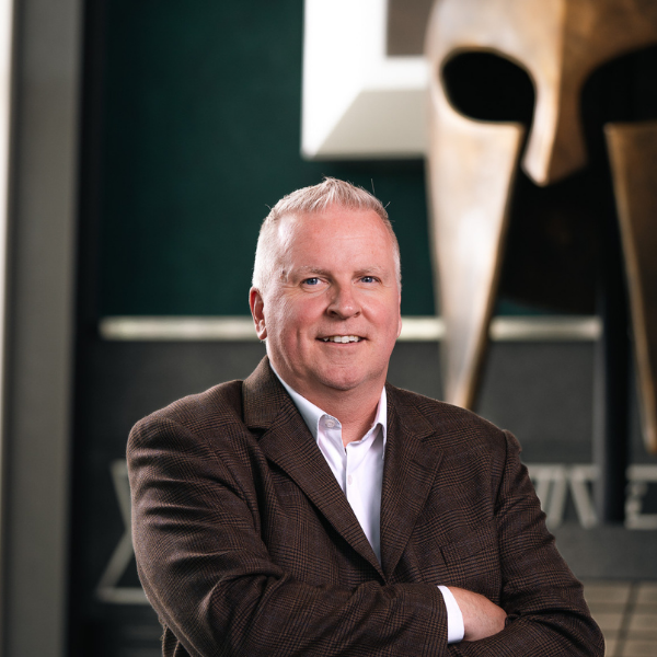 Professional headshot of a Michigan State University staff member in a patterned blazer, smiling with arms crossed in front of a Spartan helmet sculpture.