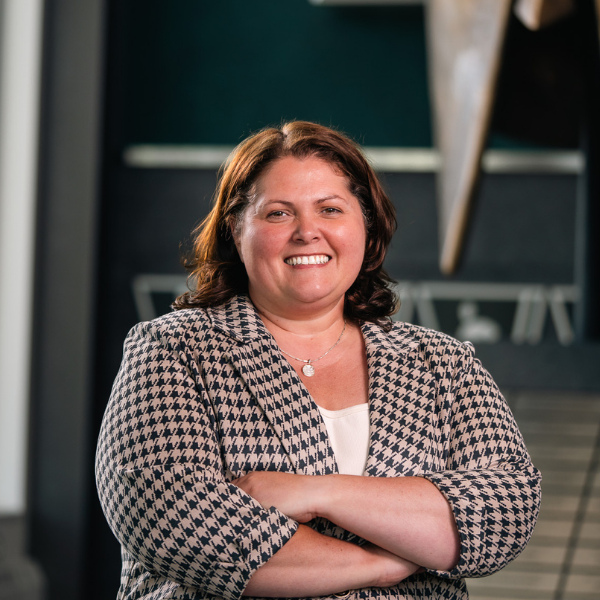 Professional headshot of a Michigan State University staff member in a patterned blazer, smiling with arms crossed in front of a Spartan helmet sculpture.