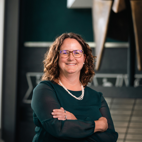 Professional headshot of a Michigan State University staff member in a green blouse and glasses, smiling with arms crossed in front of a Spartan helmet sculpture.