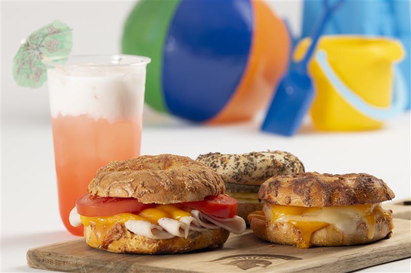 A peach colored tropical drink sits behind three bagel sandwiches oozing with cheesy eggs.