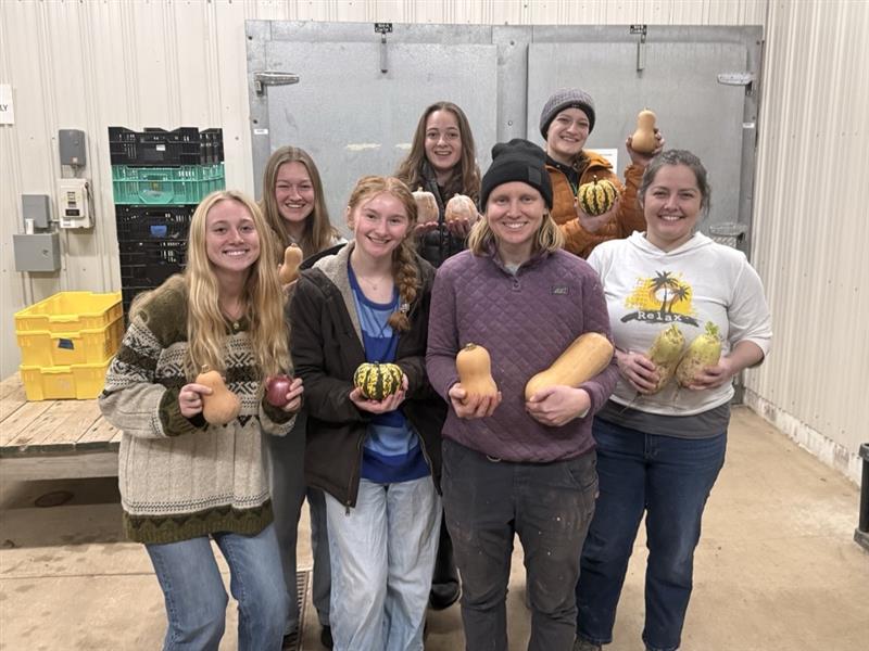 Group of students and staff smiling indoors while holding squash and gourds in a food storage or farm facility.