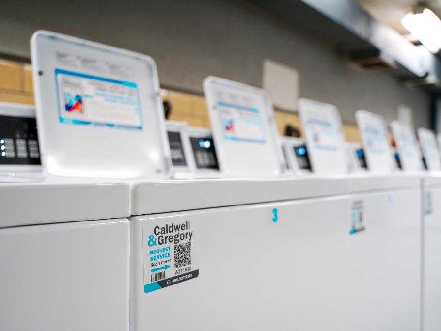 Row of new Caldwell & Gregory laundry machines in a Michigan State University residence hall, featuring ADA-accessible washers with service request QR codes.