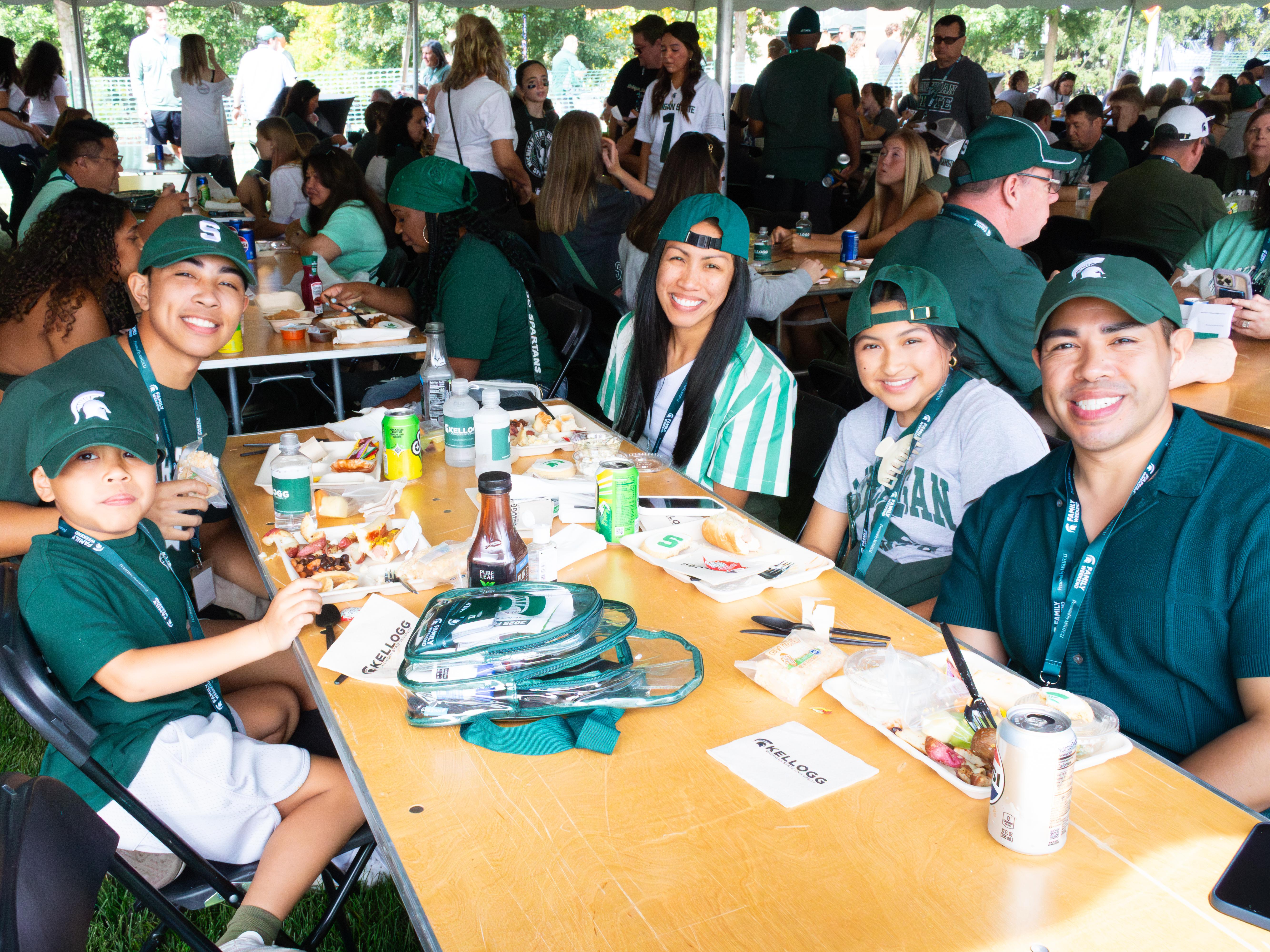Group of six people seated at a long table under an outdoor tent at a campus tailgate or gathering. The table is covered with plates of food, drinks, and event materials, including items labeled “Kellogg.” Many attendees are wearing green Michigan State University–branded shirts, hats, or lanyards. Additional tables with seated guests are visible in the background, along with trees and daylight filtering through the tent.