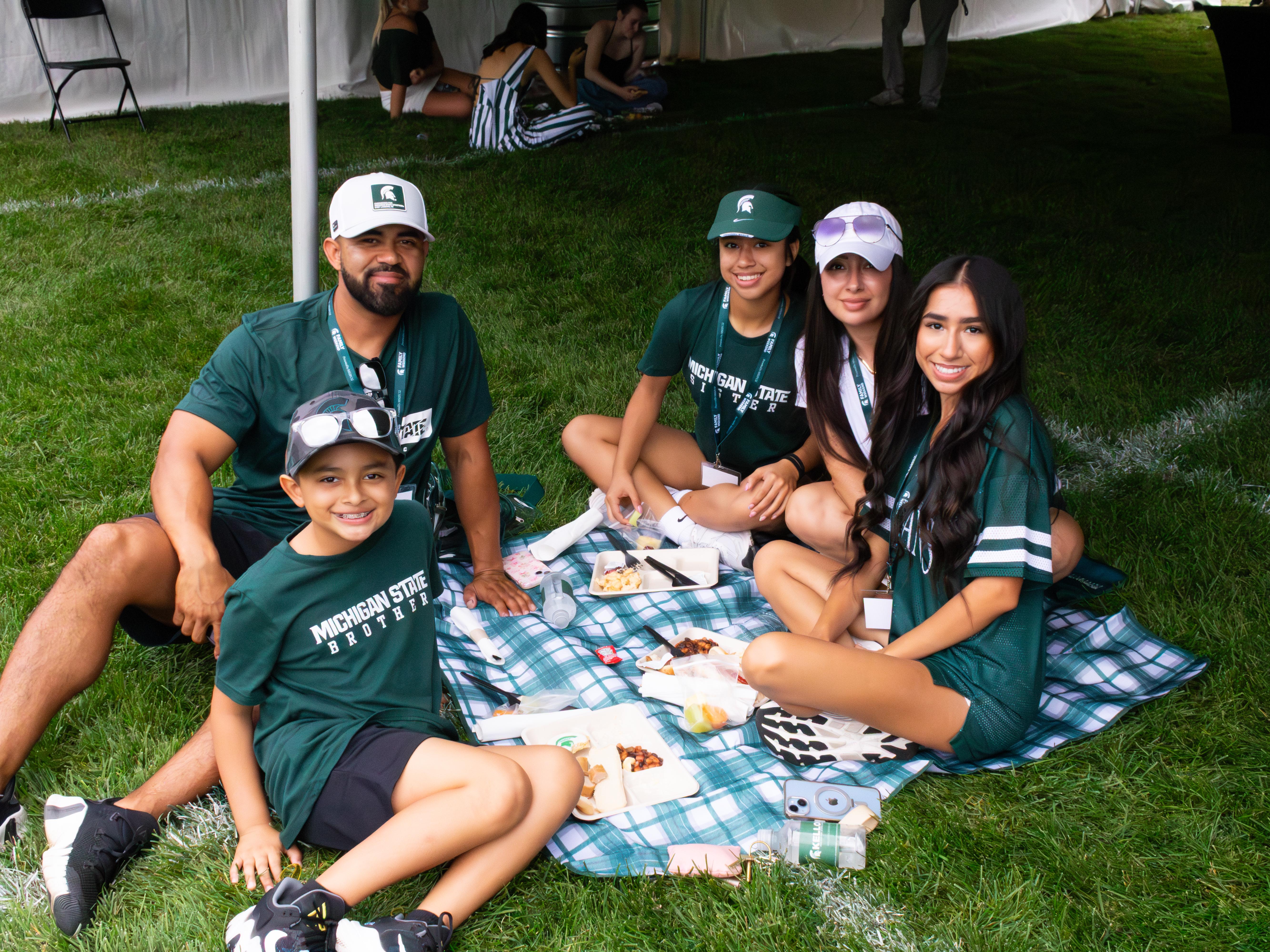 Group of six people sitting on a patterned blanket on grass at an outdoor campus event. The group is gathered under a large white tent and wearing green Michigan State University–branded shirts and hats. Plates of food and drinks are spread on the blanket, with other attendees seated and standing nearby in the background.