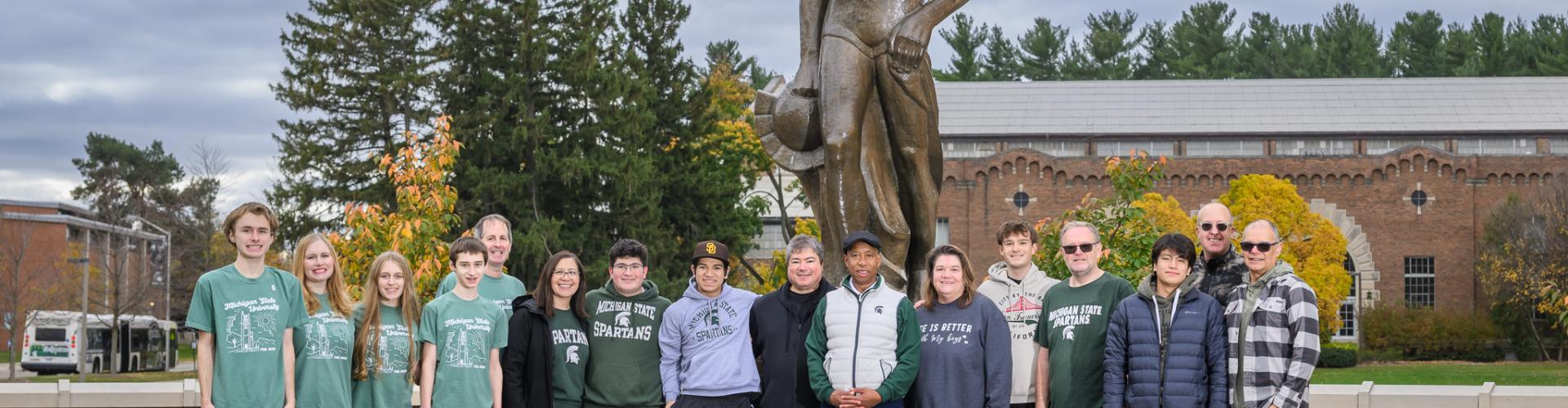 Large group of approximately fifteen people standing outdoors in front of a tall bronze statue on a brick base on a college campus. The group is arranged in a line and wearing casual, cool‑weather clothing, with many wearing green Michigan State University–branded shirts, hoodies, or jackets. Trees with autumn foliage, campus buildings, and an overcast sky are visible in the background.