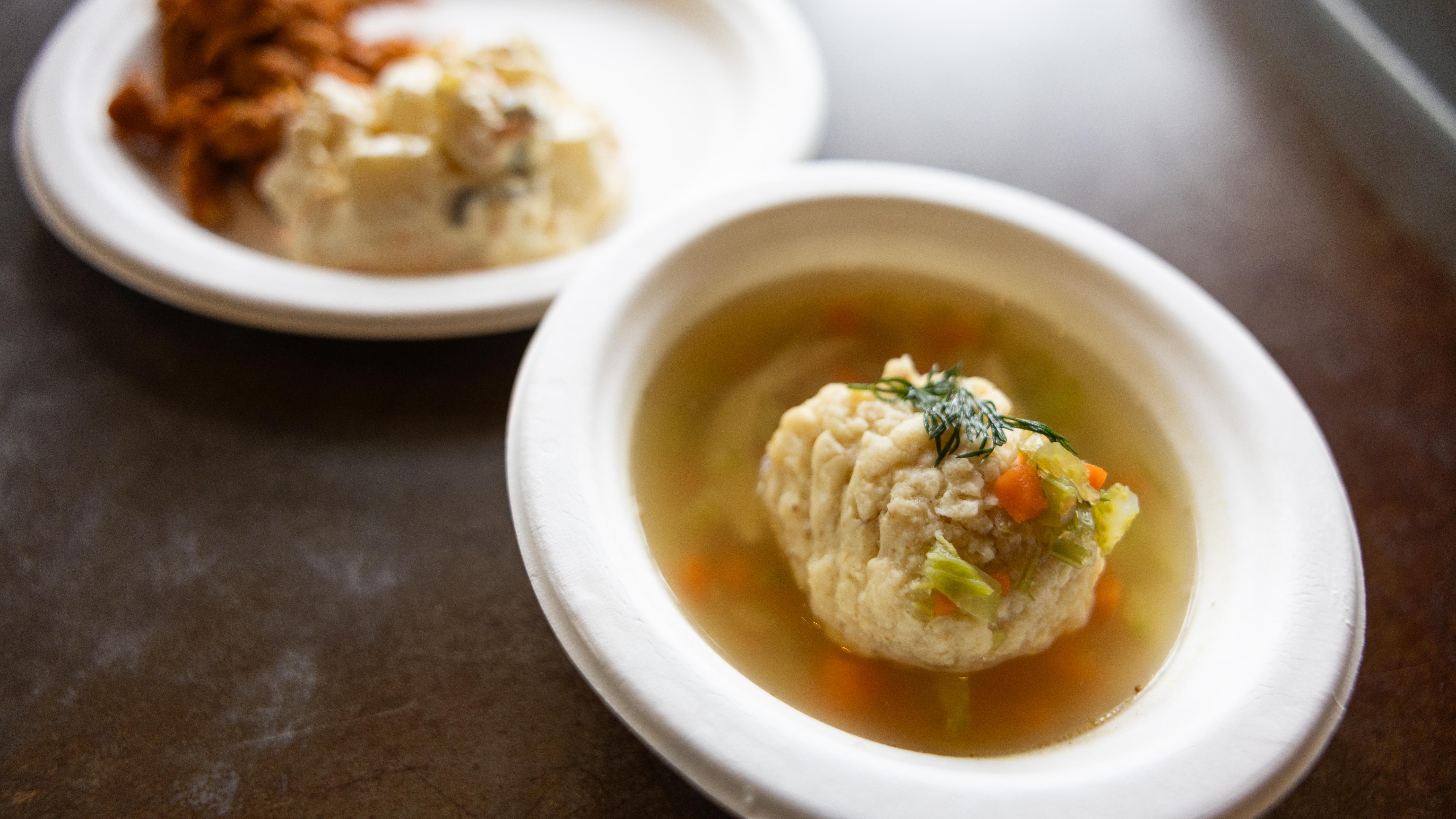 Bowl of clear soup with a large matzo ball and vegetables, with a plate of side dishes blurred in the background.