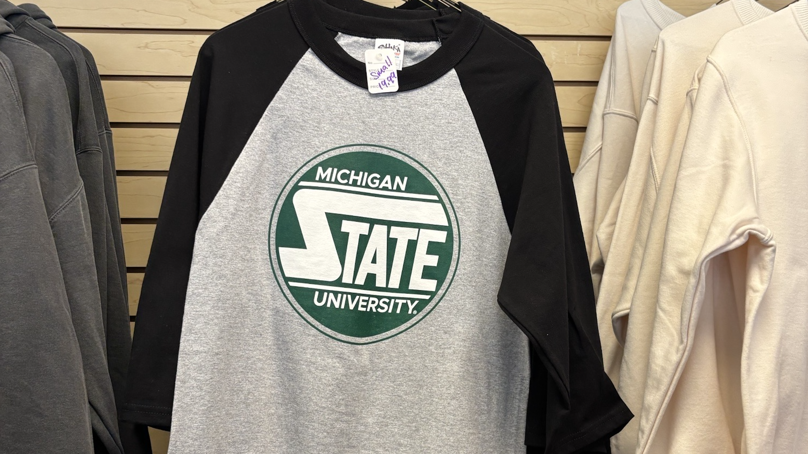 A black and gray baseball-style shirt with unique circular MSU graphic on front hangs for sale at Spartan Linen Services.