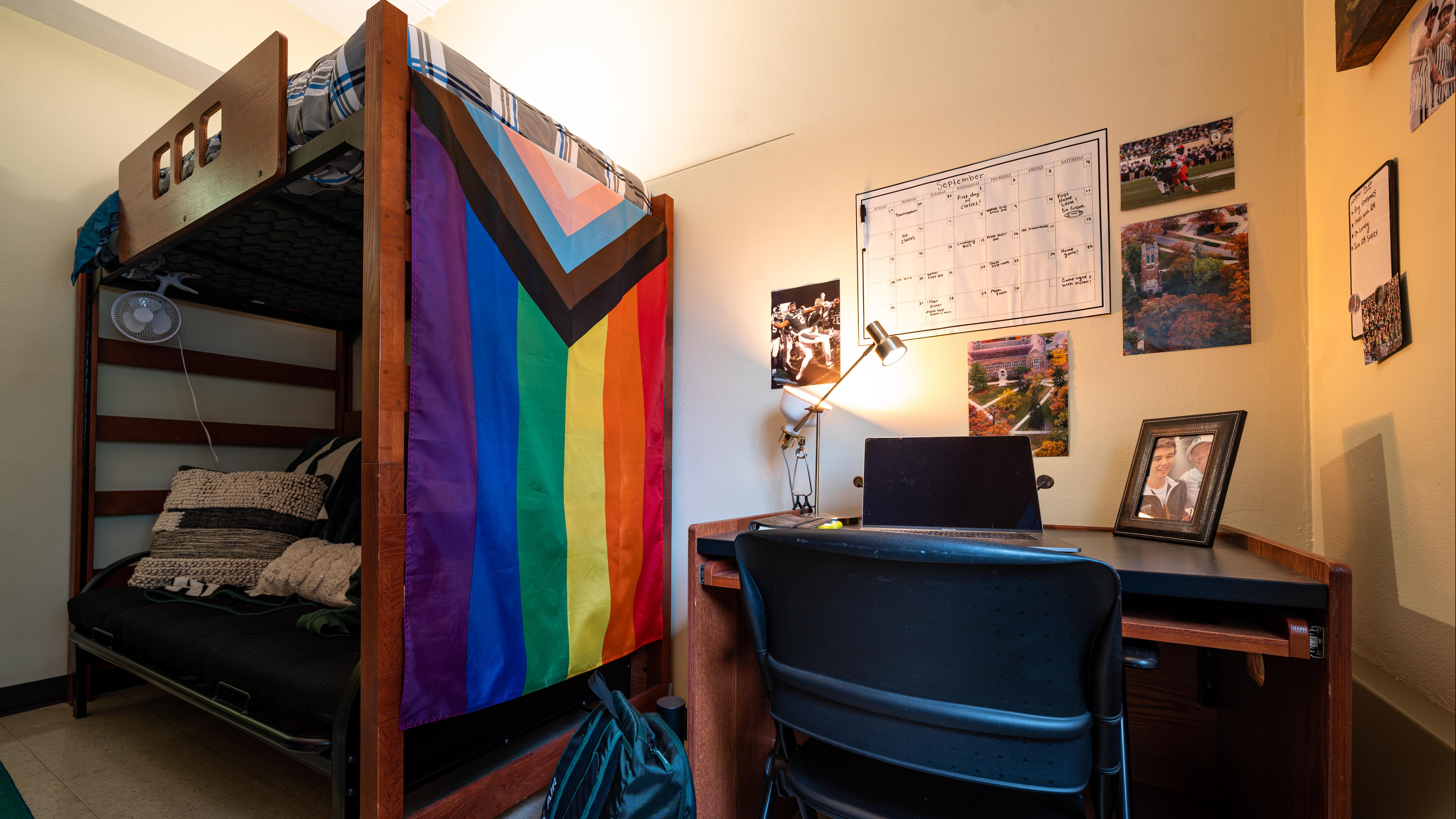 A warmly lit college dorm room with a wooden lofted bunk bed on the left and a desk on the right. A large Progress Pride flag hangs from the side of the bed above a cushioned futon. The desk holds an open laptop, adjustable lamp, and framed photo, with a black chair in front and a backpack on the floor. A monthly calendar, campus photographs, and other personal pictures decorate the walls.