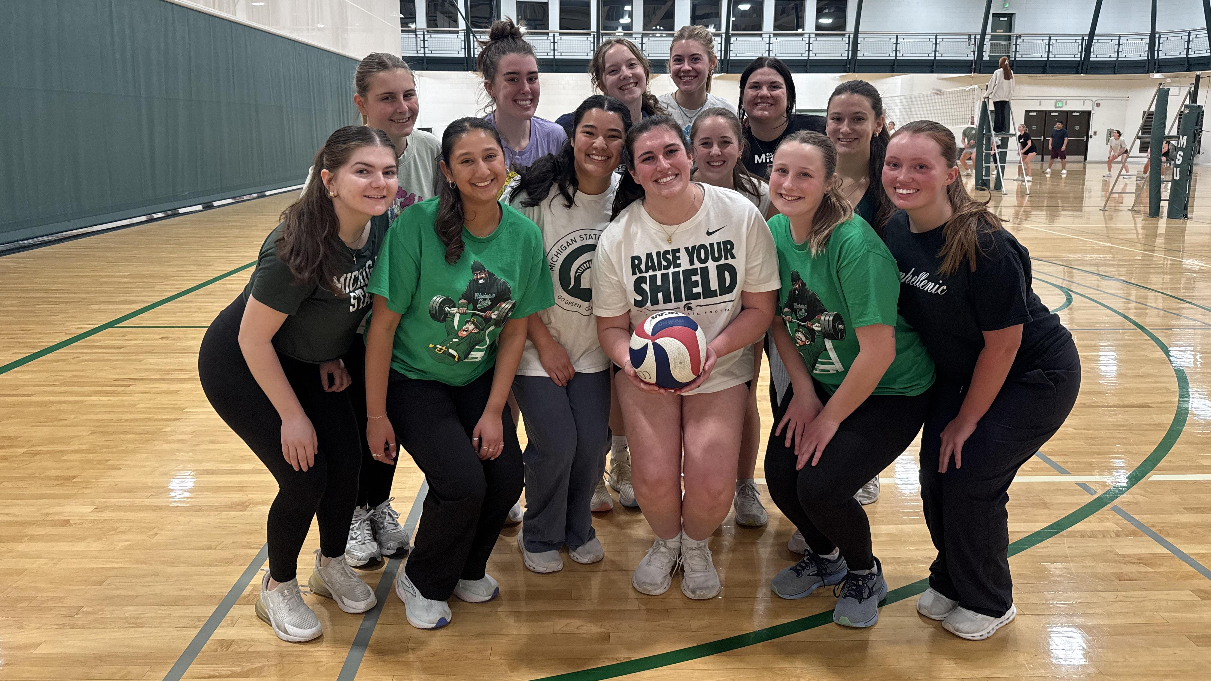 A group of 13 people pose together on a polished indoor volleyball court, smiling toward the camera. The person in the center holds a red, white, and blue volleyball and wears a “Raise Your Shield” T-shirt. Several others wear green Michigan State–themed shirts. Volleyball nets, basketball hoops, and additional players are visible throughout the brightly lit gym.