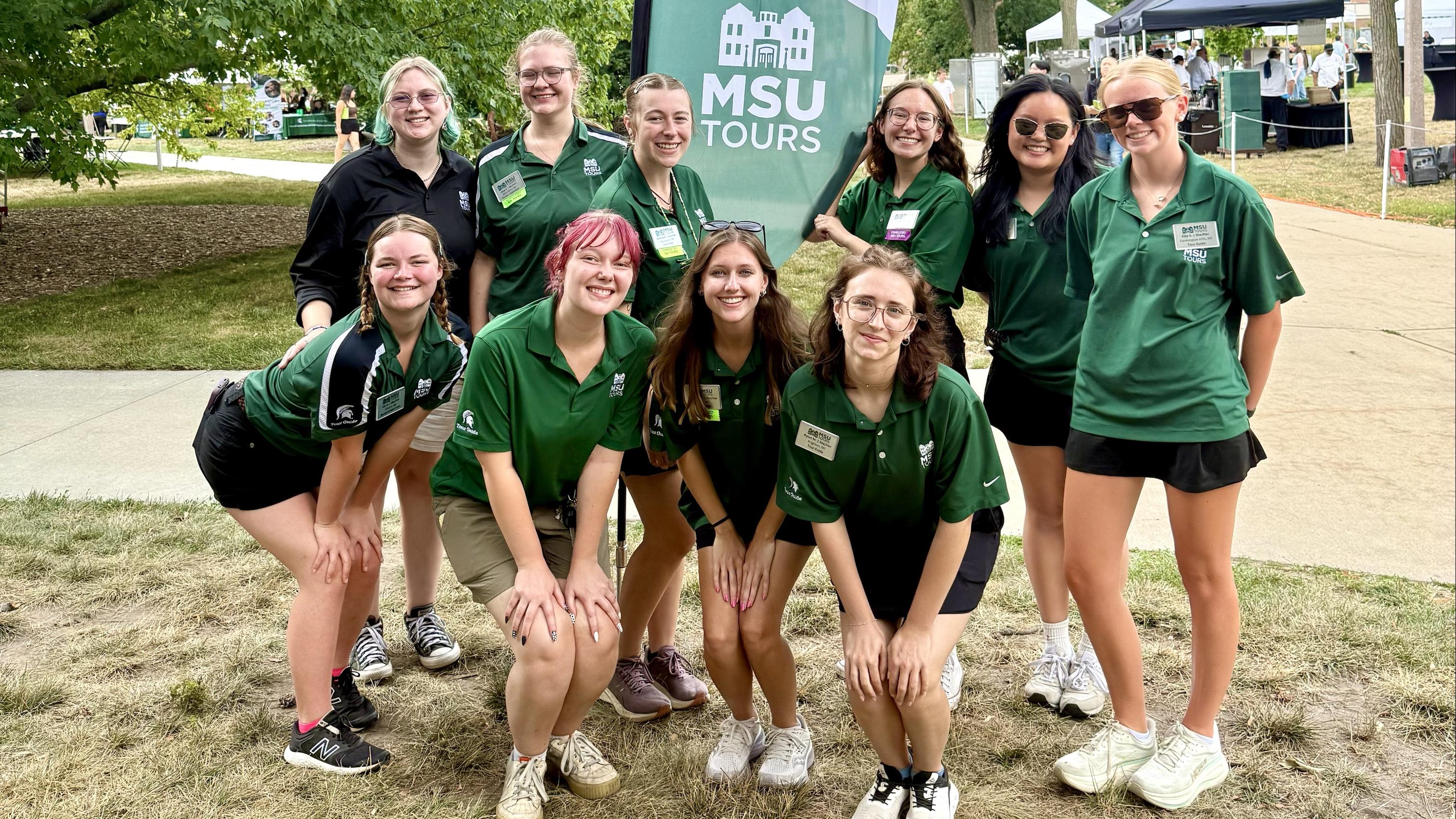 An MSU Tours group poses under a Green and White Days Tours Start Here banner on campus.